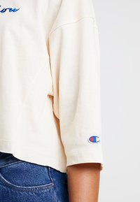 Ecrufarbener, kurzer Pullover mit lockerer Passform, der blaue gestickte Schriftzüge und ein rotes und blaues Champion-Logo am Ärmel aufweist.