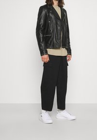 Persona che indossa una giacca di pelle nera con borchie argentate, una maglietta beige, pantaloni cargo neri a gamba larga e sneakers bianche Adidas, in piedi su un pavimento grigio.