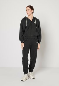 Svart huvtröja med dragsko, dragkedja och ribbade ärmar, parat med matchande sweatpants. Tyget verkar mjukt och texturerat.