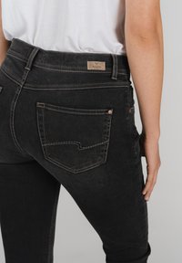 Schwarze Skinny Jeans aus Denim, mit einer Gesäßtasche mit Nähdetails und einem hellbraunen Etikett am Bund. Glatte Textur.