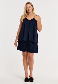 Haut sans manches en satin bleu marine avec un décolleté en V et des boutons, associé à un short assorti. Des sandales ornées de fourrure complètent la tenue.