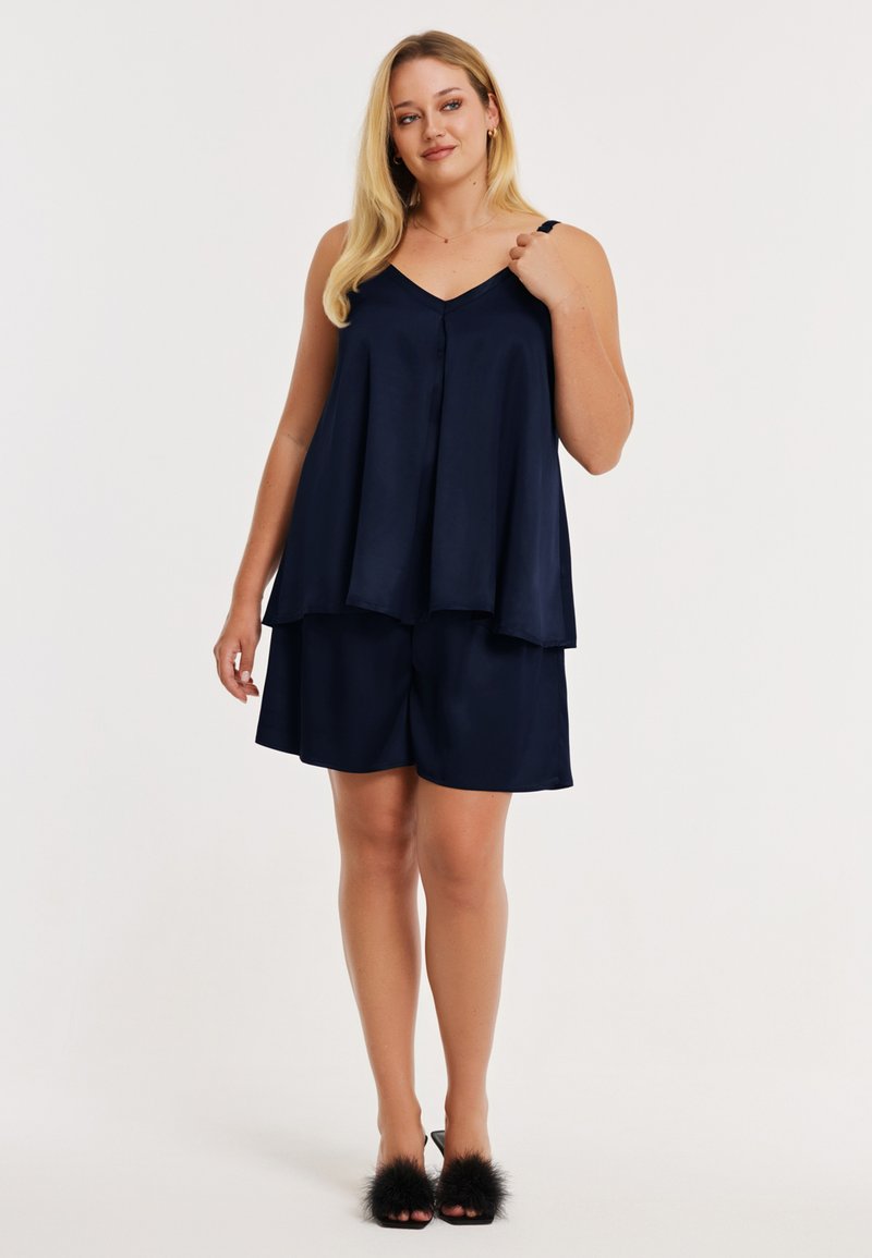 Haut sans manches en satin bleu marine avec un décolleté en V et des boutons, associé à un short assorti. Des sandales ornées de fourrure complètent la tenue.