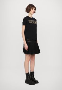 Črna majica s kratkimi rokavi z napisom "VERSACE JEANS COUTURE" v zlati barvi, v kombinaciji s črno nabrano krilo in črnimi vojnimi čevlji, ki imajo srebrne kovinske detajle.