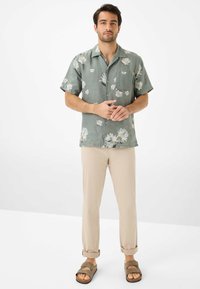 Camicetta verde chiaro a maniche corte con stampa floreale, pantaloni beige slim-fit e sandali marroni con due fasce. Tessuto morbido e traspirante.