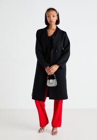 Black wool coat with double-breasted buttons, worn over a black top and red wide-leg pants. Pair of silver strappy heels and a small handbag.
