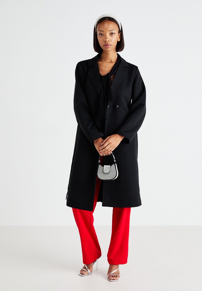 Cappotto di lana nero con bottoni doppi, indossato sopra un top nero e pantaloni rossi a gamba larga. Un paio di décolleté argentate con cinturini e una piccola borsa a tracolla.