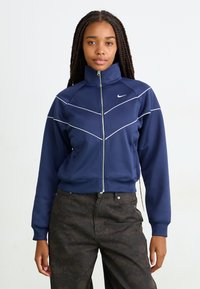 Temno moder zip-up jopič z visokim ovratnikom, okrašen z belimi diagonalnimi šivi in majhnim logotipom. Narejen iz gladkega, elastičnega materiala.