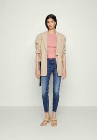 Blazer bege sobre uma blusa rosa, combinado com jeans skinny azuis e sandálias de tiras cor de canela. O blazer apresenta um único botão e mangas descontraídas.