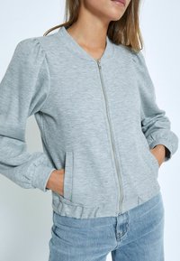 Šedá bomber bunda na zips vyrobená z textúrovanej látky má rebrované manžety, okrúhly výstrih a bočné vrecká. Rúcha sú jemne zhabané.