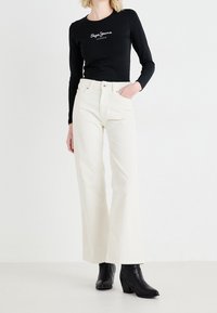 Woman wearing a black long-sleeve top with "Pepe Jeans London" logo, white high-waisted wide-leg jeans, and black ankle boots.