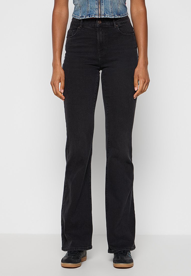 Femme portant un jean bootcut noir taille haute, des baskets noires et un haut en denim bleu sans manches avec zip, se tenant contre un fond blanc.