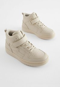 Baskets montantes beige en matériau synthétique lisse, avec un col rembourré, un devant lacé et une sangle velcro ajustable.