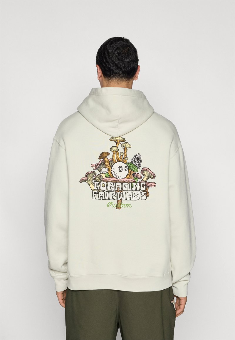 Personne portant un sweat à capuche beige clair avec un motif coloré de champignon et le texte "Foraging Fairways" dans le dos, associé à un pantalon vert olive.
