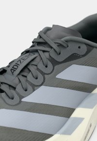 Zapatilla deportiva gris con parte superior texturizada, que presenta un diseño de rayas gris y azul claro, logo prominente y cordones duraderos.