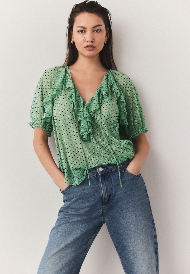 Green polka dot blouse featuring ruffled accents, a V-neckline, and short sleeves, paired with high-waisted blue denim jeans.