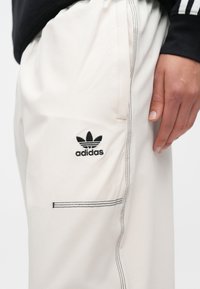 Αθλητικό παντελόνι ανοιχτού μπεζ χρώματος με μαύρη ραφή και πλαϊνή τσέπη, με κεντημένο μαύρο λογότυπο Adidas �στο αριστερό μηρό.