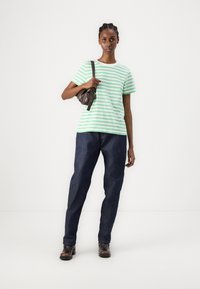 Grønn og hvit stripete bomull t-skjorte, avslappet passform, sammen med mørkeblå denim jeans og brune skinn ankelsko. Modellen holder en liten brun veske.