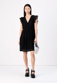 Vestido preto em camadas com detalhes de ilhós, decote em V e mangas com babados; combinado com uma clutch cinza claro e sapatos plataforma pretos e robustos.