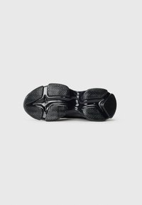Semelle de sneaker noire avec un motif de tread texturé, comprenant des grips en zigzag et une arche centrale lisse, sur un fond clair uni.