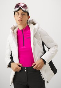 Weißer, puffy Jacke mit Kapuze, die ein schwarzes Seitenpanel hat. Darunter ein leuchtend pinkes langärmliges Oberteil mit einem schwarzen Reißverschluss. Schwarze Skihose.