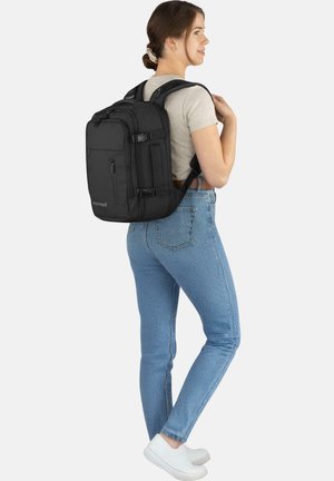 Black fabric backpack with front zip pocket and side straps. Model wearing light gray shirt and blue jeans, standing from a side view.