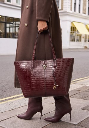 Osoba nosząca brązowy płaszcz i buty na obcasie, trzymająca dużą ciemno bordową torbę typu tote ze skóry tłoczonej w wzór krokodyla z złotym breloczkiem w kształcie serca.
