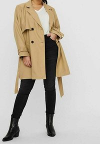 Beige Trenchcoat mit großen Revers, doppelt geknöpft und mit einem Bindegürtel; kombiniert mit schwarzen Skinny Jeans und schwarzen Stiefeletten.