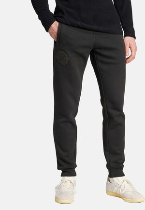 Pantalones deportivos negros hechos de una tela suave, con un diseño ajustado y un parche de logo en relieve en el muslo izquierdo. Combinados con zapatillas ligeras.
