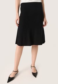 Črna pletena krilo s širokim robom, gladke teksture, srednje dolžine in oprijetim pasom. Nosi se z črnimi slingback čevlji s konicami.