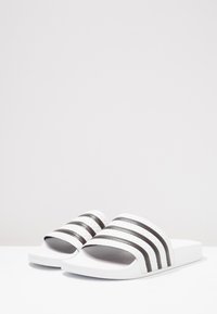 White slide sandals with three black striped accents on the upper. Features a smooth texture and a flat, contoured footbed for comfort.