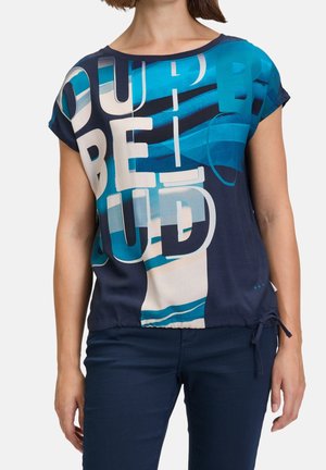 Kobieta ubrana w granatowo-turkusową bluzkę z krótkim rękawem, ozdobioną dużymi abstrakcyjnymi literami, oraz ciemnoniebieskie spodnie, stojąca na jednolitym tle.