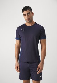 Tumed spordipaita kontrastsete siniste varrukatega, valge logo rinna peal, koos sobivate lühikesed pükstega. Sile ja veniv kangas, mis sobib spordiks.