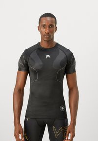 Homme athlétique portant un t-shirt de sport noir ajusté avec des motifs gris et des leggings noirs assortis avec un texte graphique jaune.