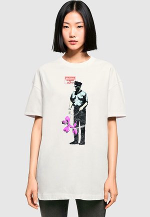 Weißes, übergroßes T-Shirt mit einem Grafikmotiv eines uniformierten Mannes, der einen rosa Ballonhund hält. Darunter steht der Text "WARNUNG WACHDUTY".