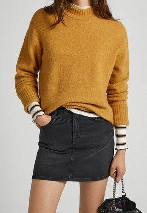 Personne portant un pull moutarde sur une chemise rayée à manches longues, mini-jupe en jean noire, tenant un sac à main noir à bandoulière chaîne.