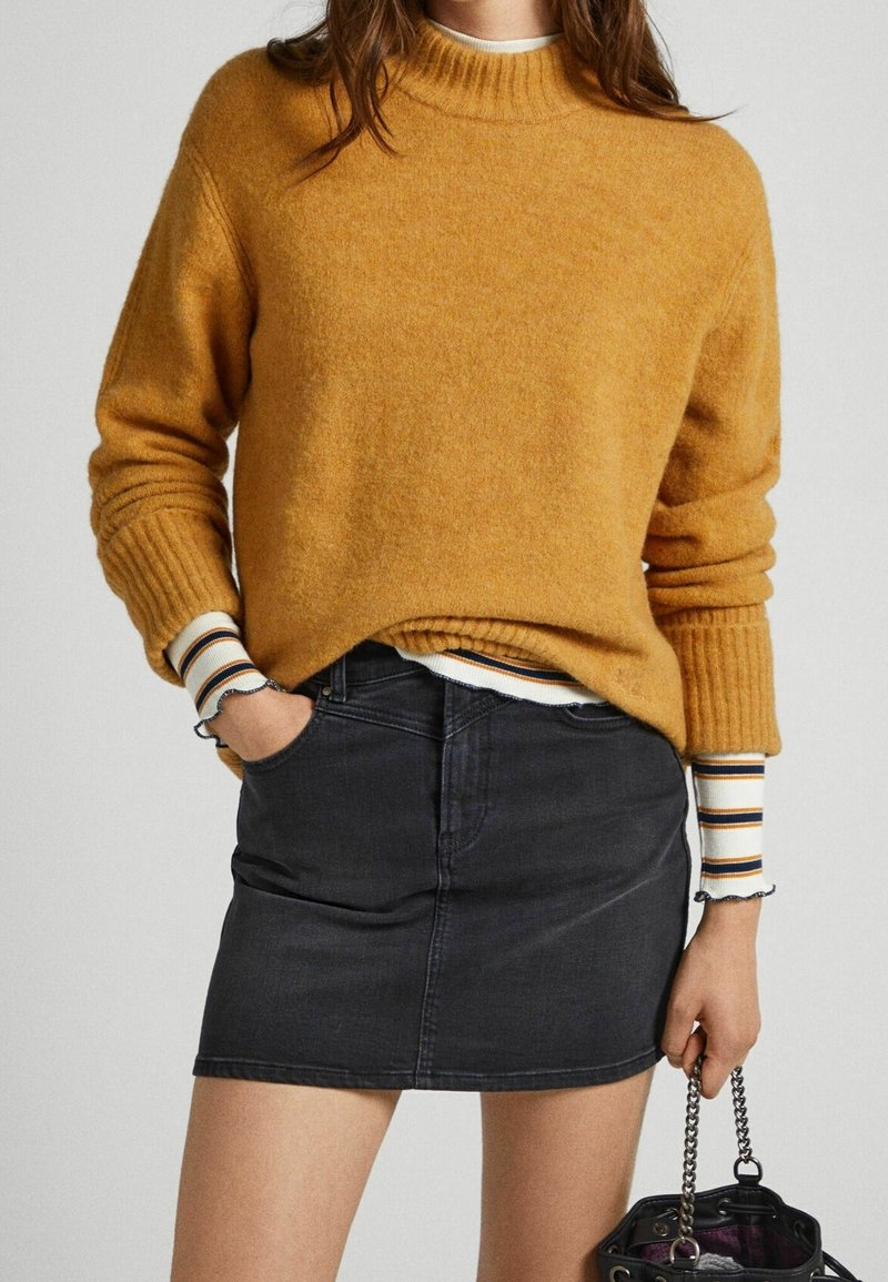 Personne portant un pull moutarde sur une chemise rayée à manches longues, mini-jupe en jean noire, tenant un sac à main noir à bandoulière chaîne.