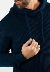 Hoodie en maille bleu foncé avec un motif texturé, doté d'une capuche avec cordon de serrage et de poignets côtelés. Main positionnée près de l'encolure.