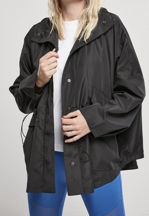 Person iført en løs sort regnjakke over en hvid skjorte og blå leggings, som lukker regnjakkens trykknapper.