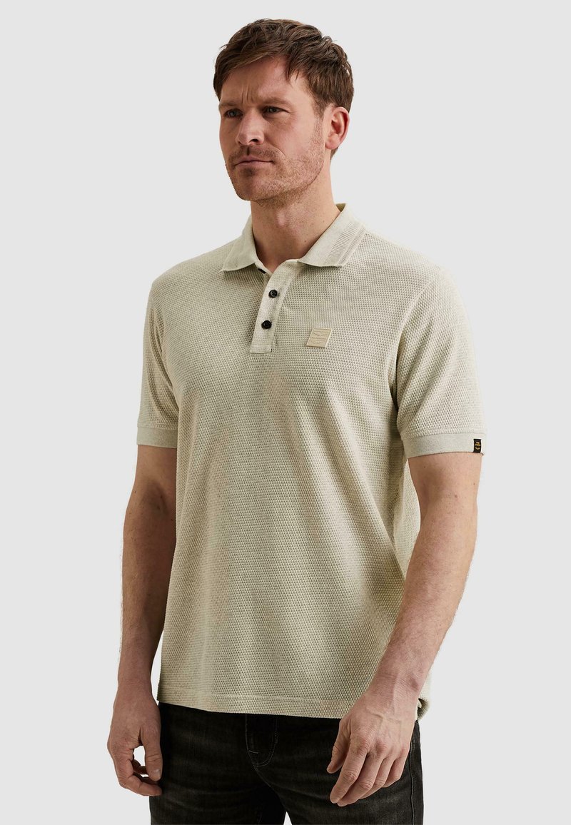 Homme portant un polo à manches courtes beige texturé avec des boutons noirs et un pantalon foncé, debout devant un fond clair uni.