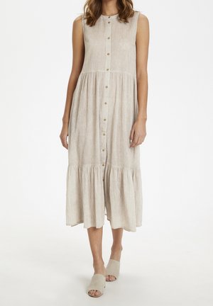 Ärmelloses, hellbeiges Midi-Kleid mit Knopfleiste und gereihten Rüschen, getragen mit passenden offenen Slip-on-Schuhen vor weißem Hintergrund.