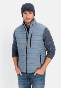 Light grey puffer vest with horizontal stitching, black zippered pockets, and a brown leather accent, paired with a dark knit beanie.