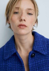 Modra teksturirana jakna s ovratnikom in črnim gumbom za zapiranje. Posnetek obraza osebe z kratkimi lasmi in nevtralnim make-upom.