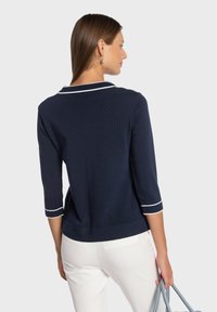 Pull marine avec bordure blanche, texture côtelée, manches trois-quarts et col rond. Porté avec un pantalon blanc et un sac à main bleu clair.