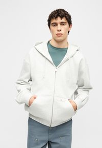 Νέος άνδρας που φοράει ελαφρύ γκρι φούτερ με φερμουάρ πάνω από πράσινο πουκάμισο, με τα χέρια στις μπροστινές τσέπες, στέκεται μπροστά σε ένα απλό λευκό φόντο.