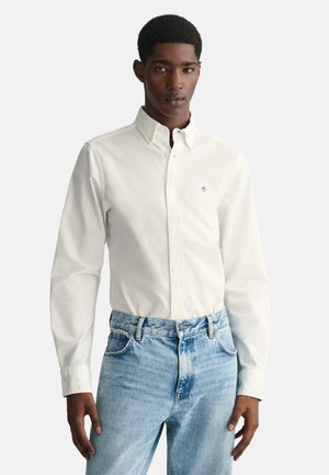 Camisa blanca de botones con cuello clásico y mangas largas, combinada con jeans de mezclilla azul claro. Tejido texturizado con sutil detalle de logo.