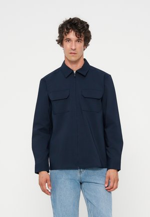 Homme aux cheveux bouclés portant une veste zippée bleu marine avec deux poches poitrine et un jean bleu clair, debout devant un fond blanc uni.