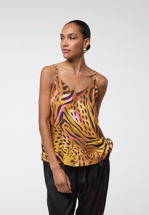 Mujer con el cabello recogido en un moño, que lleva pendientes de aro dorados, una camiseta sin mangas con estampado de rayas de tigre en amarillo, rosa y negro, y pantalones negros.