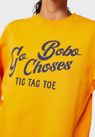 Felpa gialla brillante con testo blu navy che legge "Go Bobo Choses TIC TAC TOE." Presenta un collo rotondo e maniche lunghe.