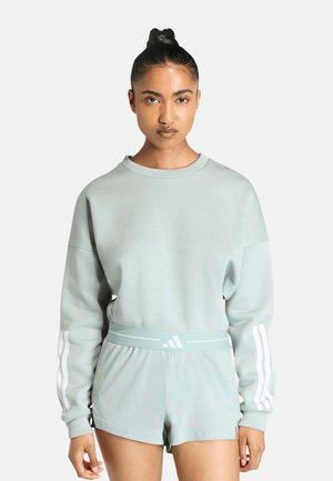 Femme portant un sweat à manches longues bleu clair et un short assorti avec des bandes blanches Adidas sur les manches et un logo à la taille, debout face à nous.