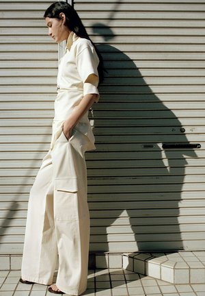 Kobieta w kremowym stroju i czarnych sandałach stoi bokiem z rękami w kieszeniach przy zamkniętej metalowej rolce, rzucając długi cień.